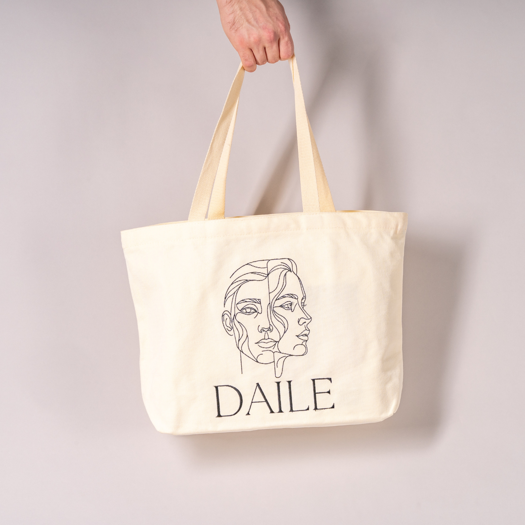Beige canvas tote bag with abstract face line art and 'DAILE' text, hand holding handle