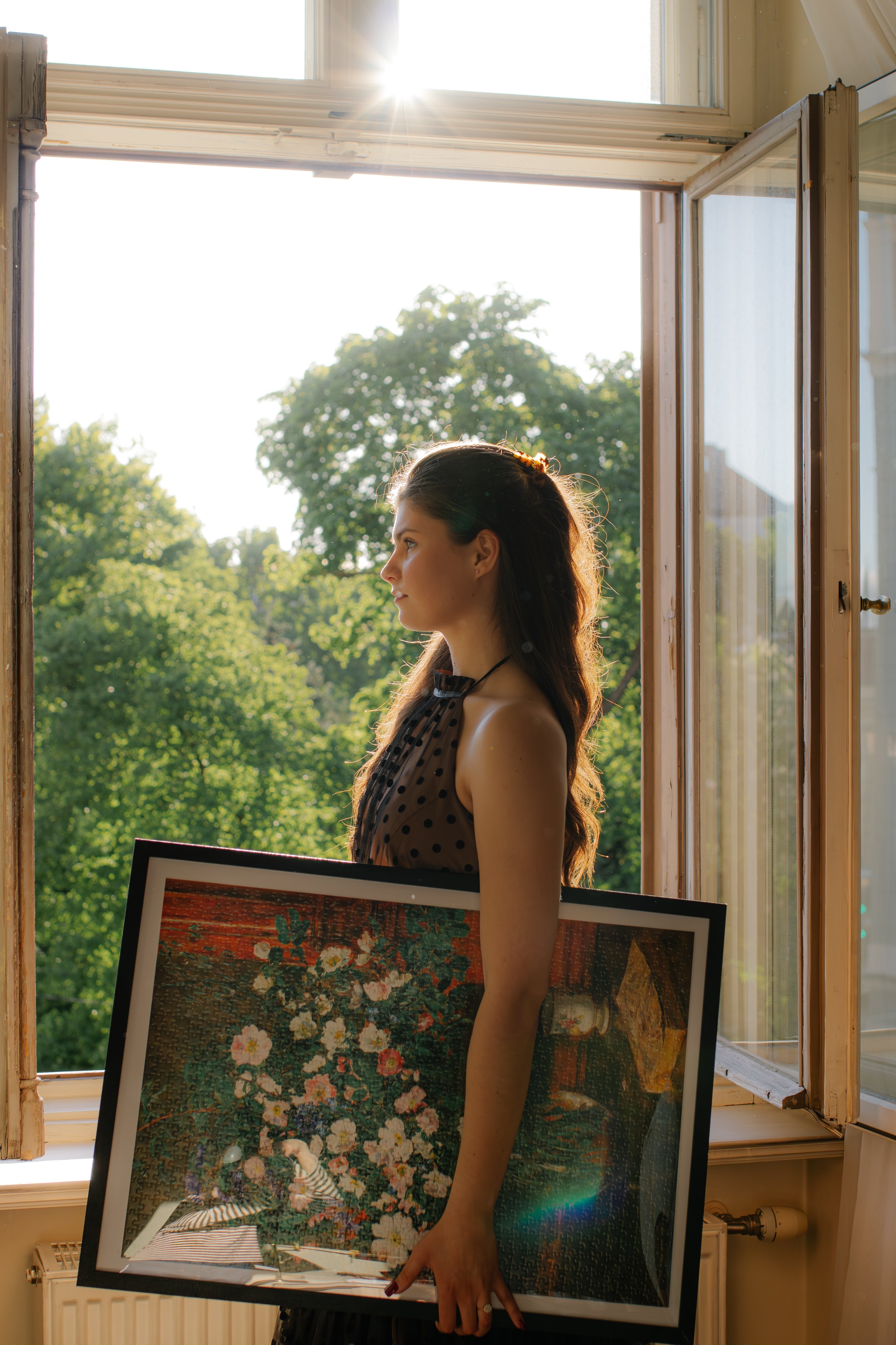 Woman holding floral art puzzle by window, museum-quality collectible with sunlit garden view