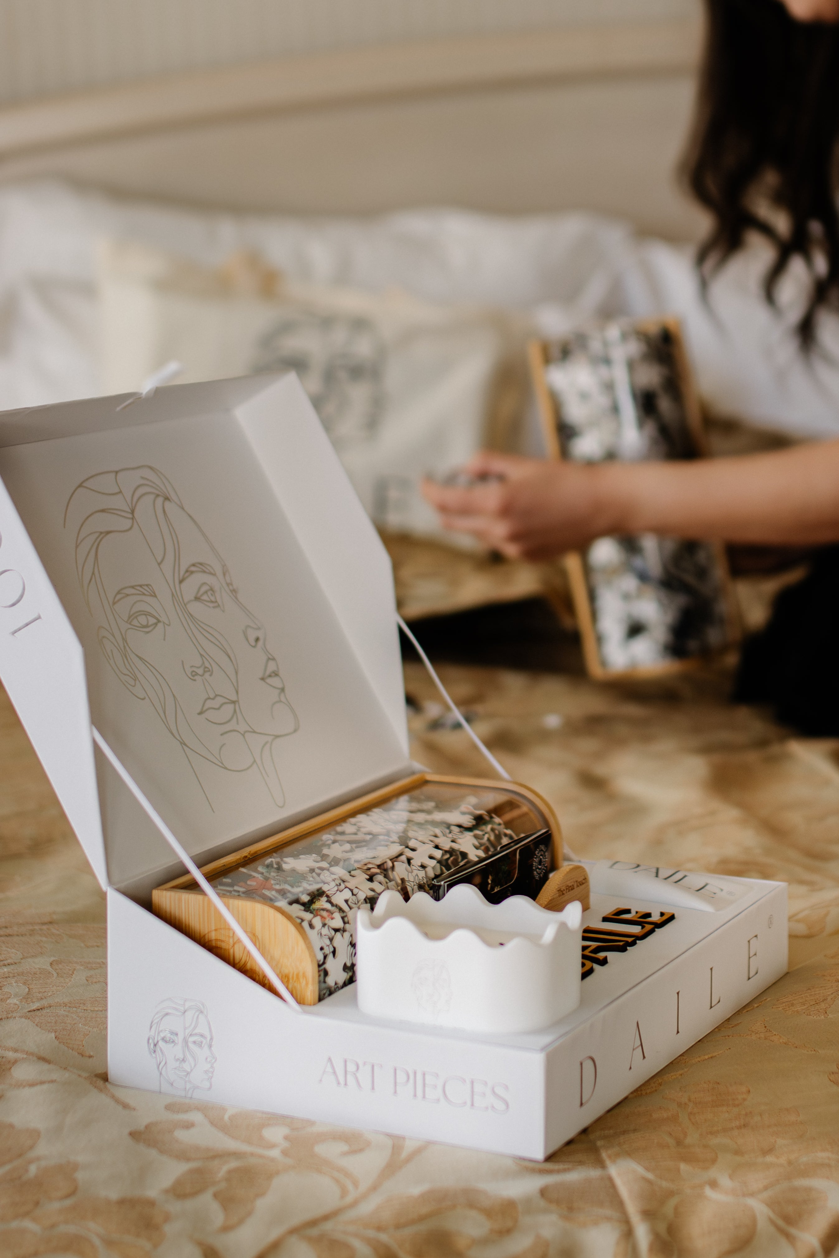Premium art puzzle box with line art portrait, wooden tray, and puzzle pieces, styled on a bed for cozy unboxing.
